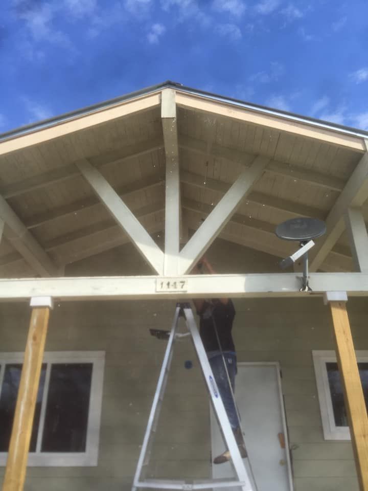Ladder leaned against a house porch roof with white trusses under a blue sky.