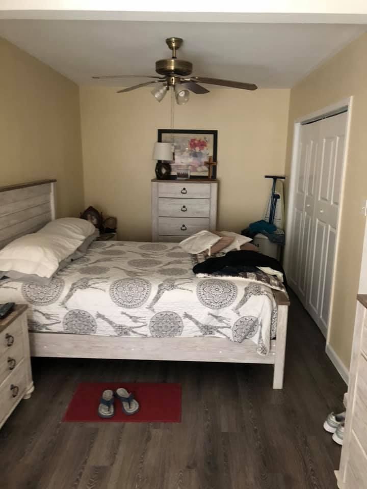 Small bedroom with a made bed, dresser, ceiling fan, closet, and shoes on a red mat.