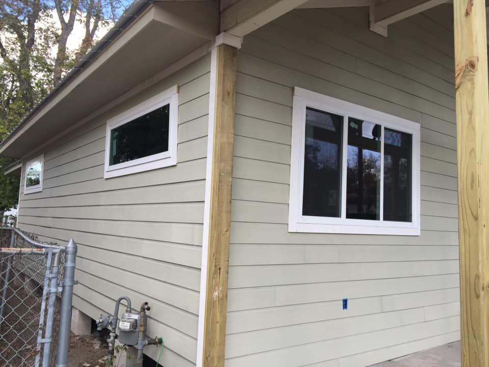 Side of a beige house with white-trimmed windows and exposed wooden corner posts near a chain-link fence.