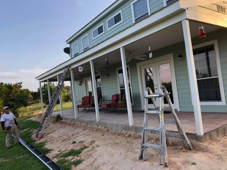 Workers on a porch with ladders, tools, and soil grading beside a light blue house with white columns