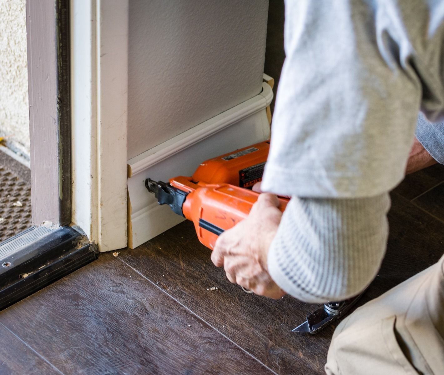 Person using an orange power tool to drill near a door frame on a dark floor indoors