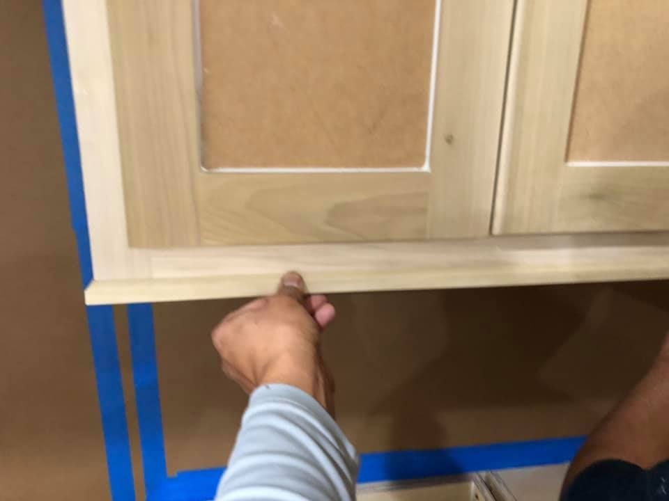 Hands installing a light wood cabinet under a counter, with blue painter’s tape on the wall and floor.