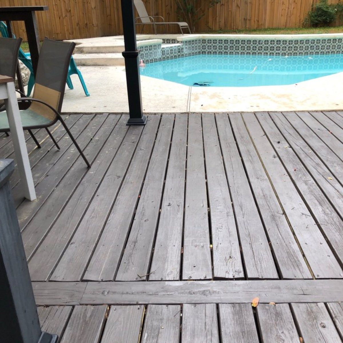 Wooden deck beside a backyard pool with patio chairs and a black umbrella pole