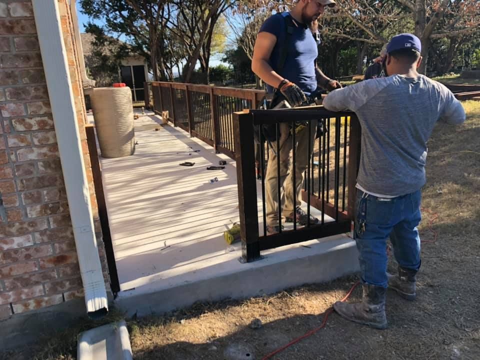 Two workers installing a black railing on a wooden ramp beside a brick building
