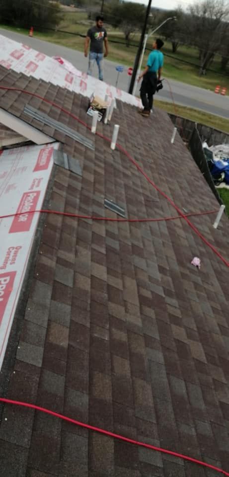 Workers on a steep shingled roof, installing red safety lines and materials near a road.