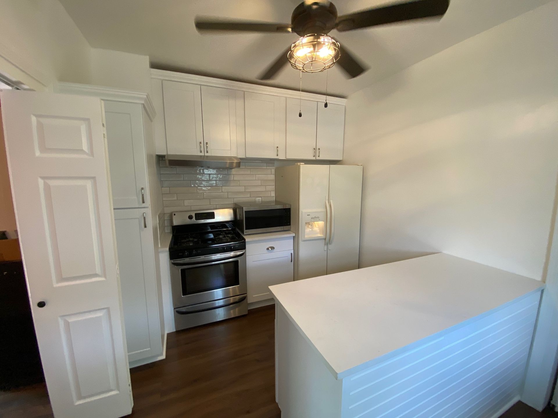 Small white kitchen with stainless stove and fridge, white cabinets, ceiling fan, and hardwood floor.