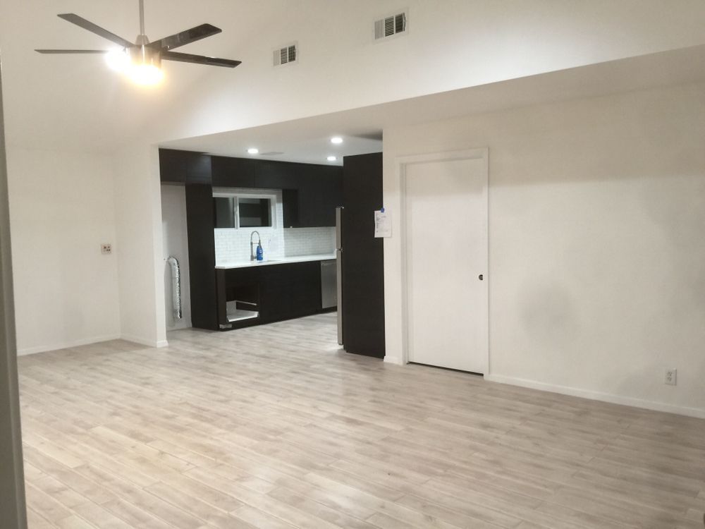 Empty modern living room with light wood floors, ceiling fan, and open kitchen in the background