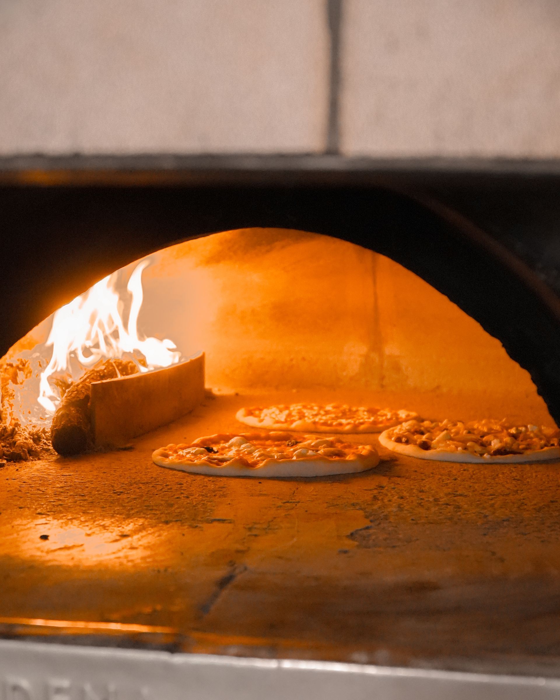 Pizza in cottura in un forno a legna, con le fiamme a sinistra e due pizze sulla pietra rovente.