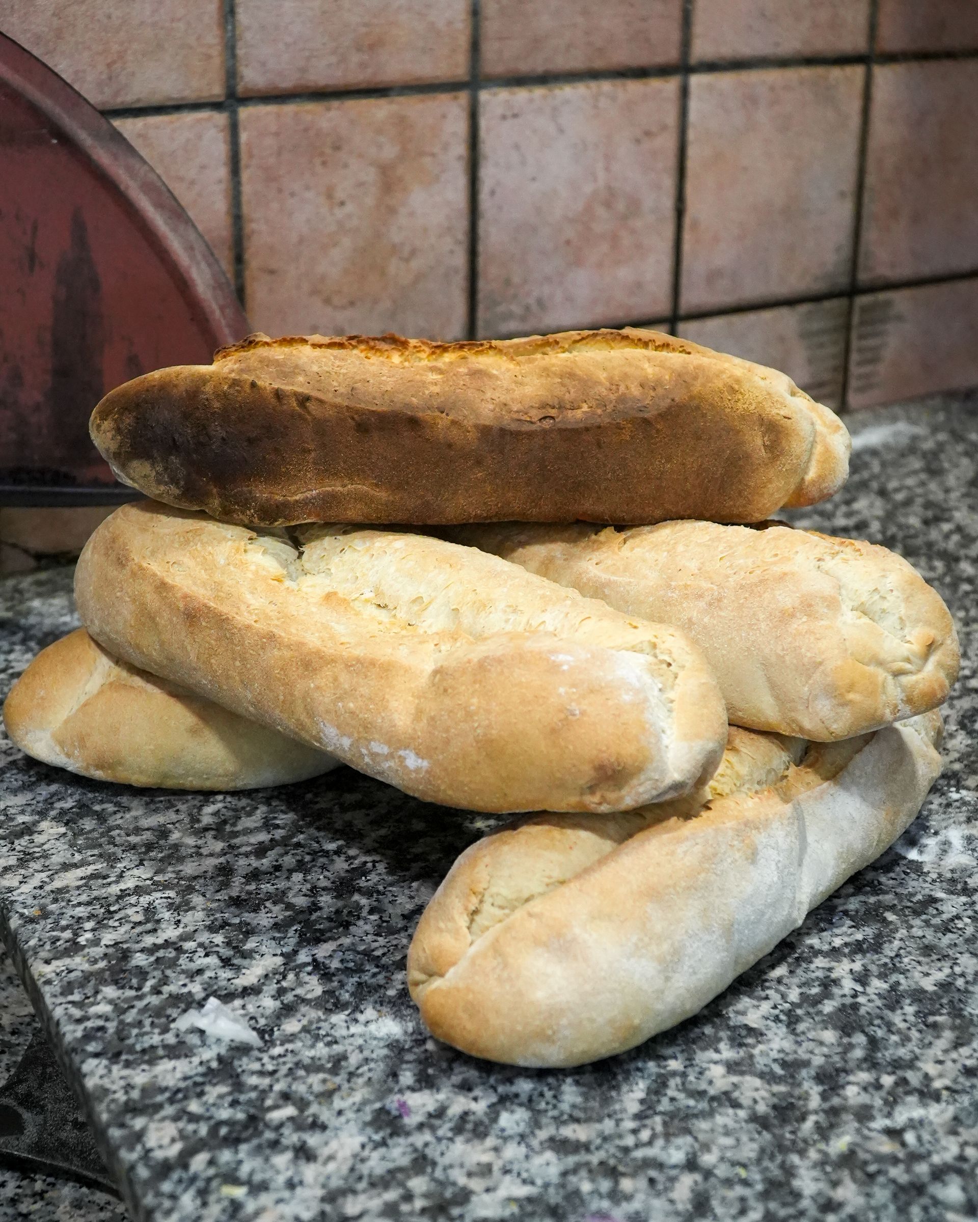 Diverse pagnotte di pane impilate su un piano di lavoro in granito, con una pagnotta scura e ben cotta in cima.