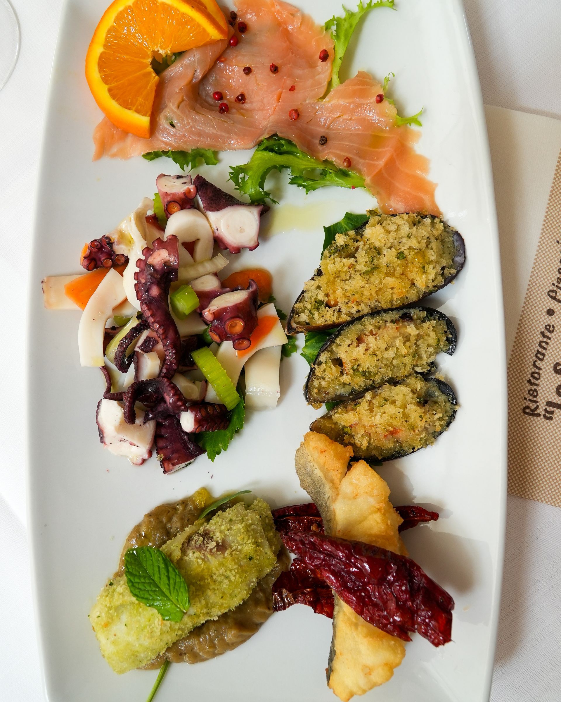 Assorted gourmet appetizers on a white plate, including salmon, citrus, salad, stuffed mussels, and mashed greens.