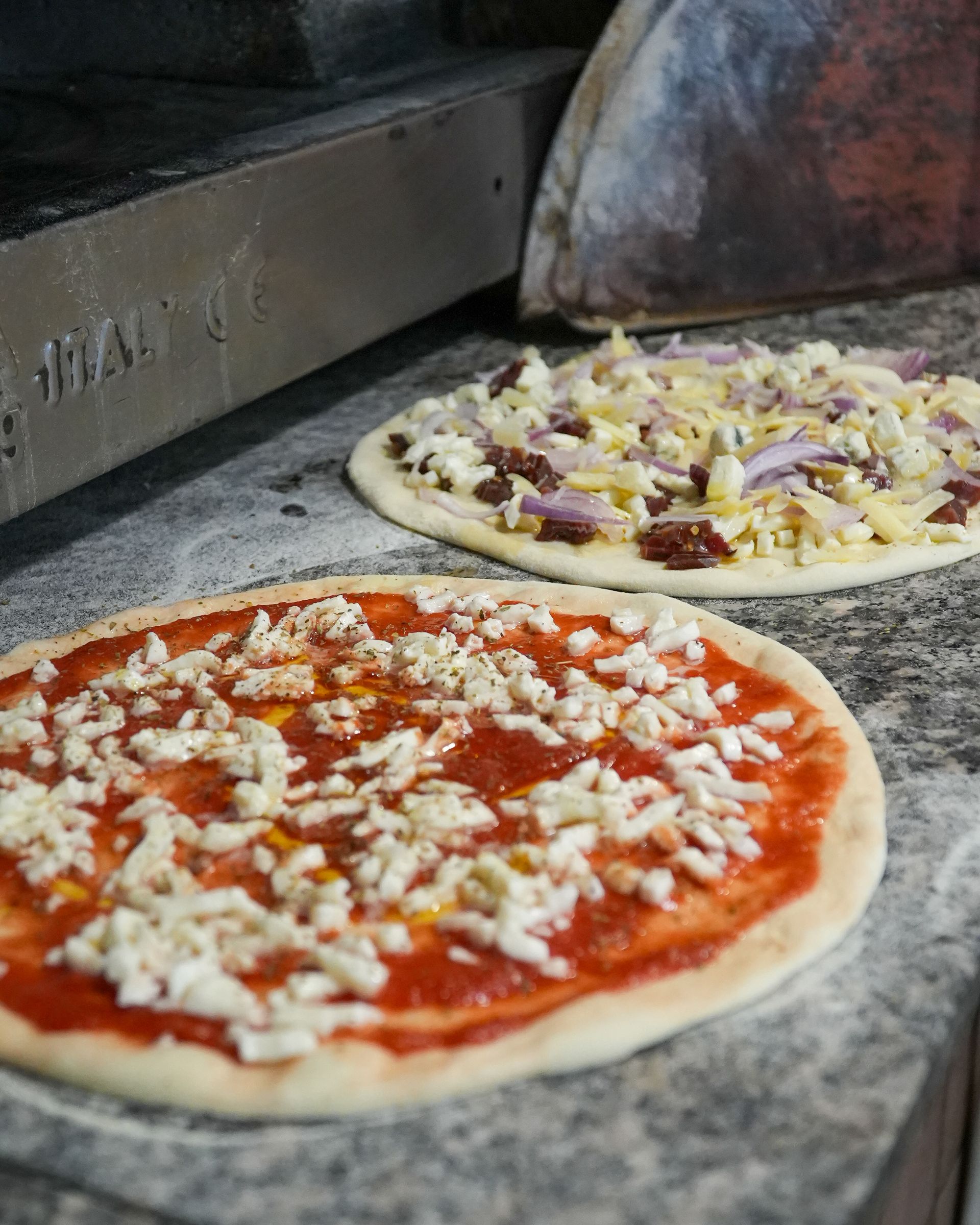 Due pizze crude su un bancone di pietra, una con salsa di pomodoro e formaggio, l'altra con i condimenti, che vengono infornate.