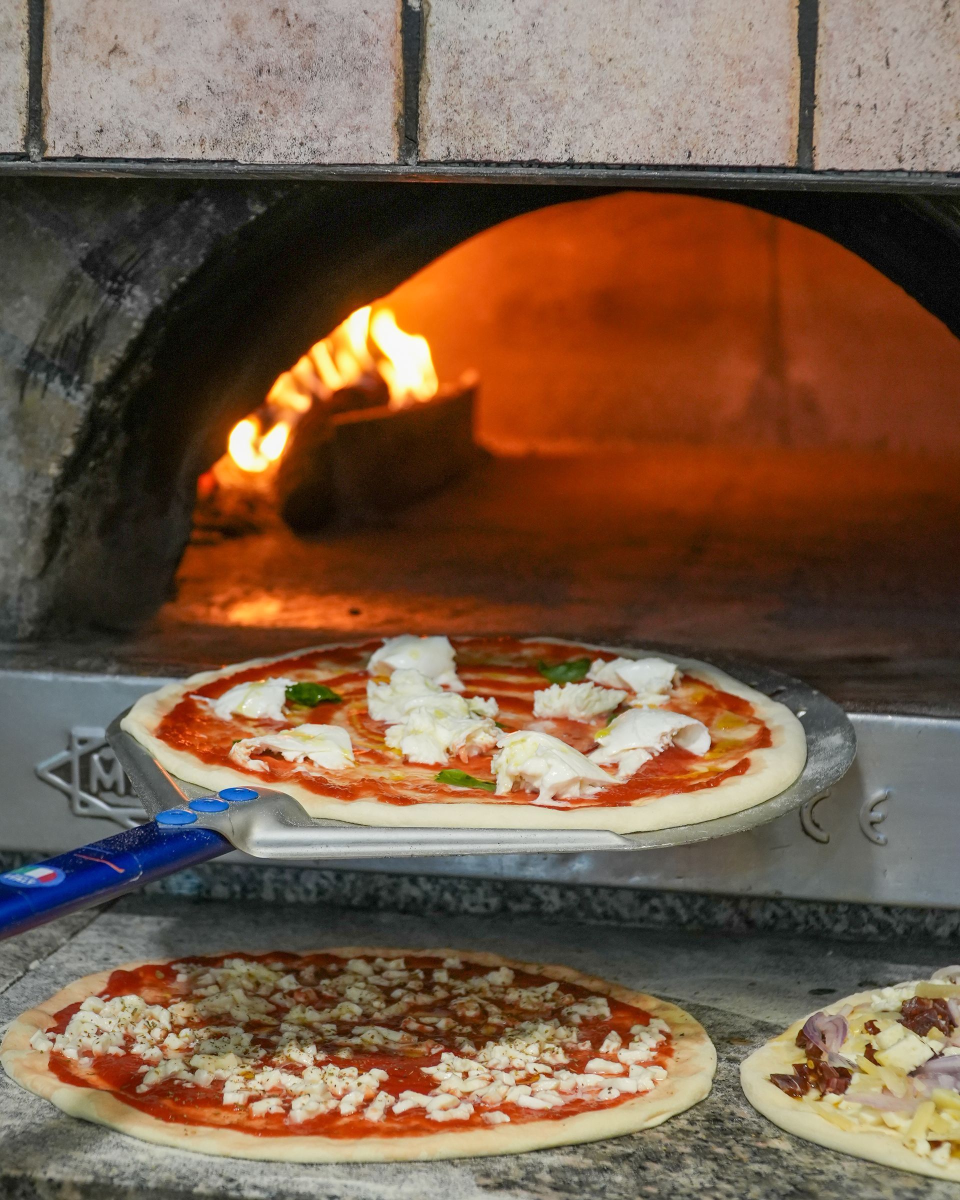 Pizza in cottura in un forno a legna, con due pizze pronte su delle teglie davanti.