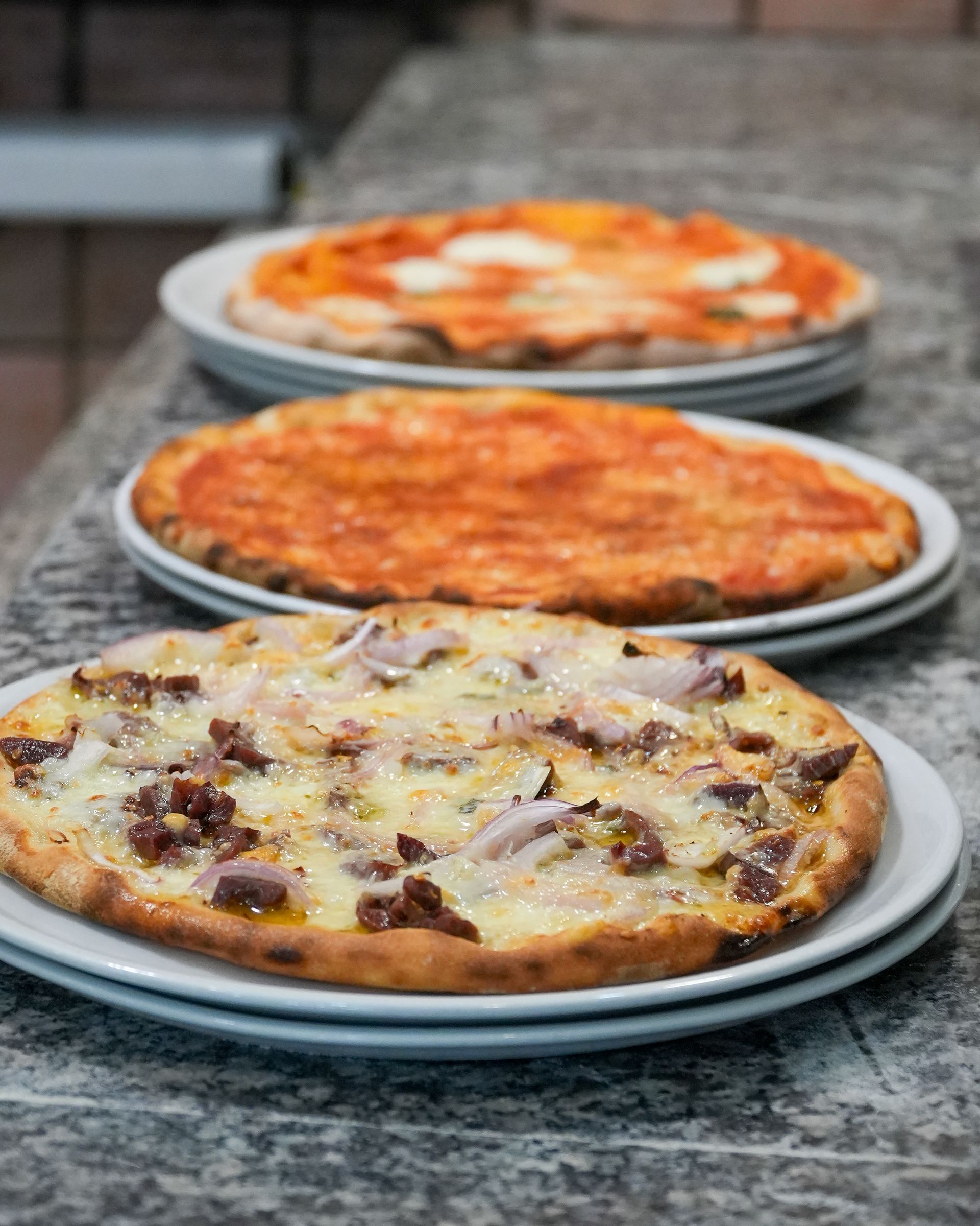 Tre pizze su piatti appoggiati su un piano di lavoro in granito, tra cui una condita con funghi e salsiccia.