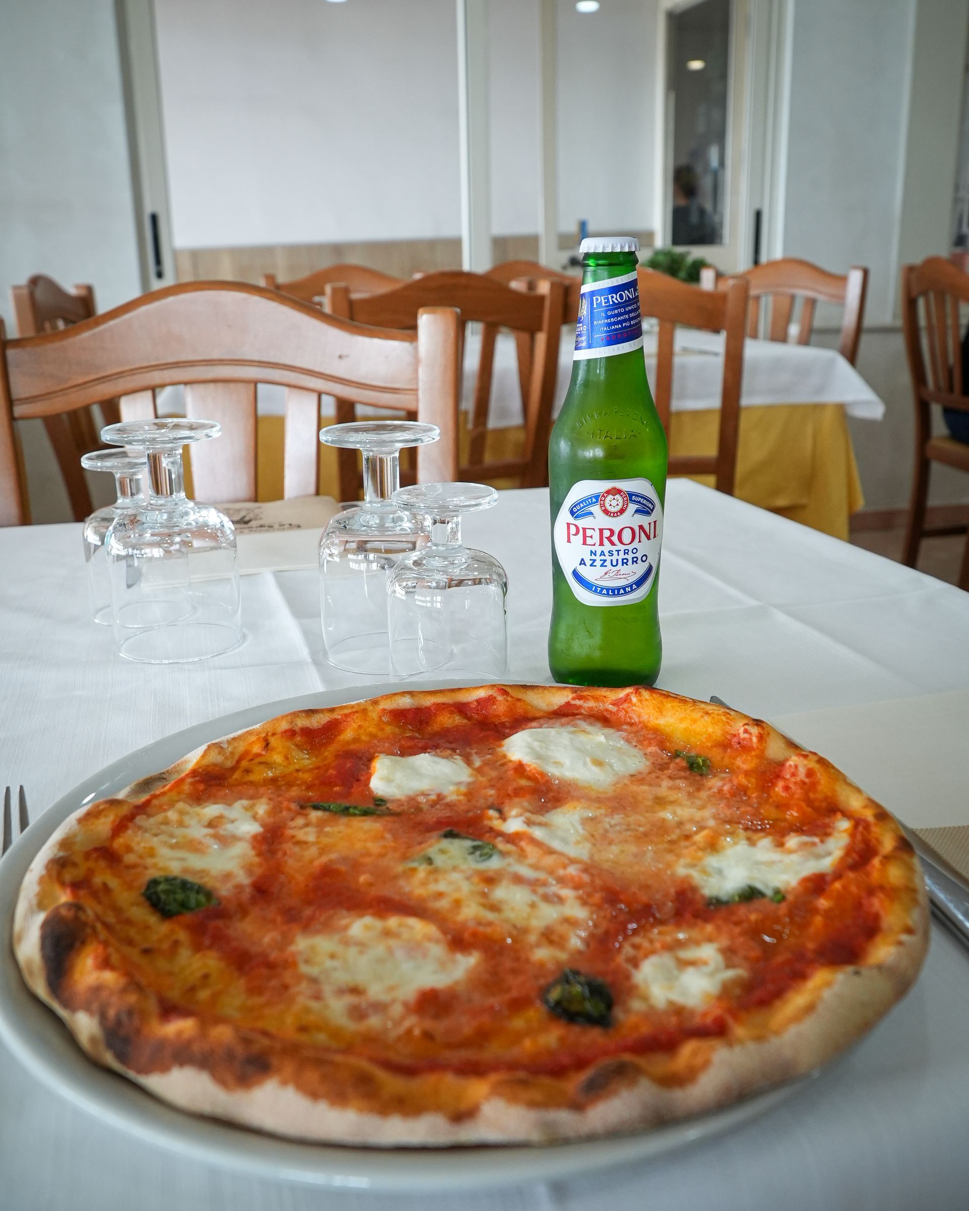 Pizza e bottiglia di birra verde su un tavolo di ristorante, con bicchieri e sedie sullo sfondo.