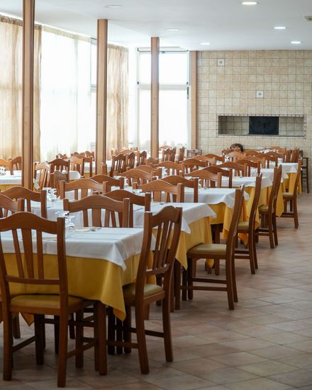 Sala da pranzo vuota di un ristorante con sedie di legno, tovaglie bianche e grandi finestre.