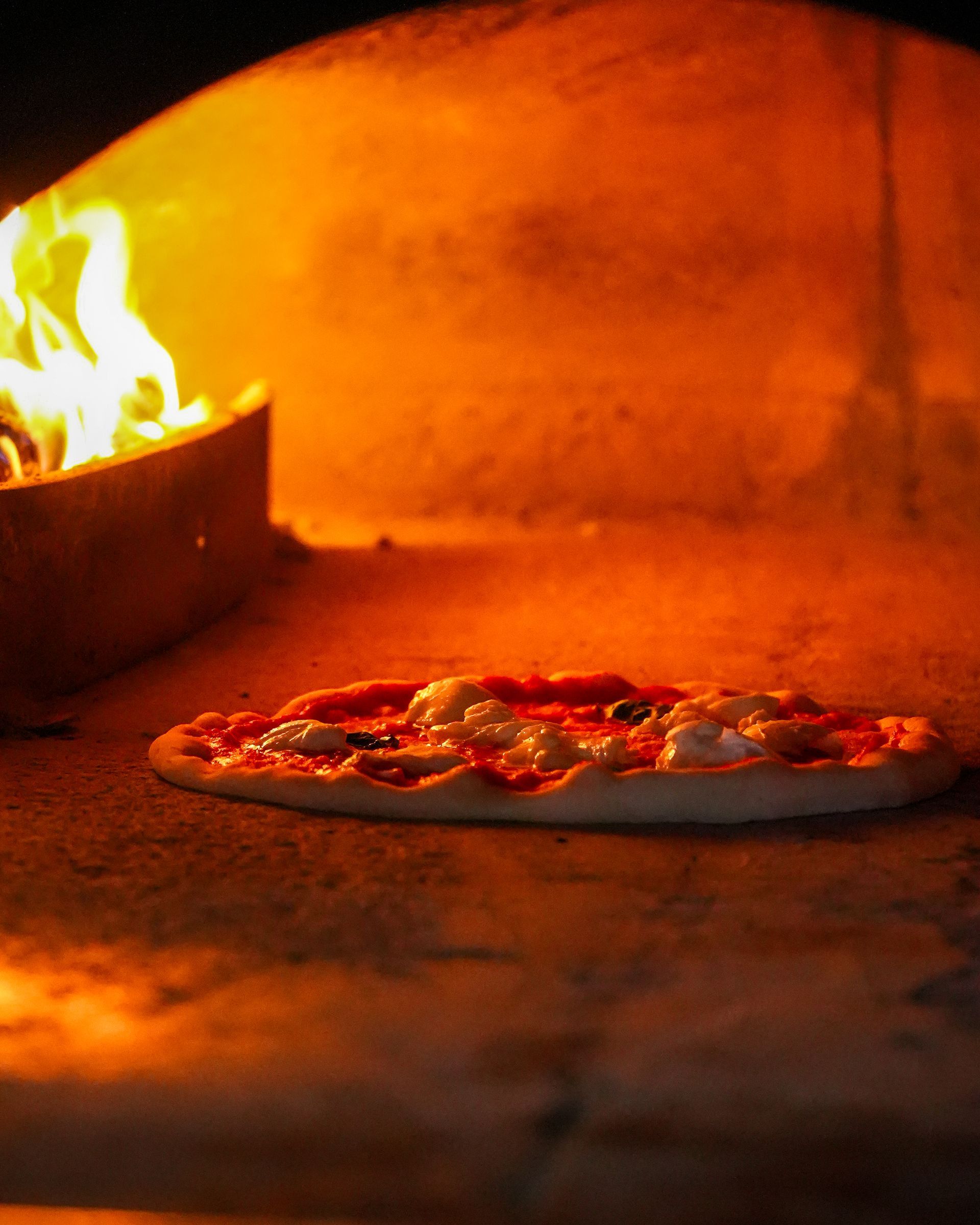 Pizza in cottura in un forno a legna con le fiamme che brillano sulla sinistra