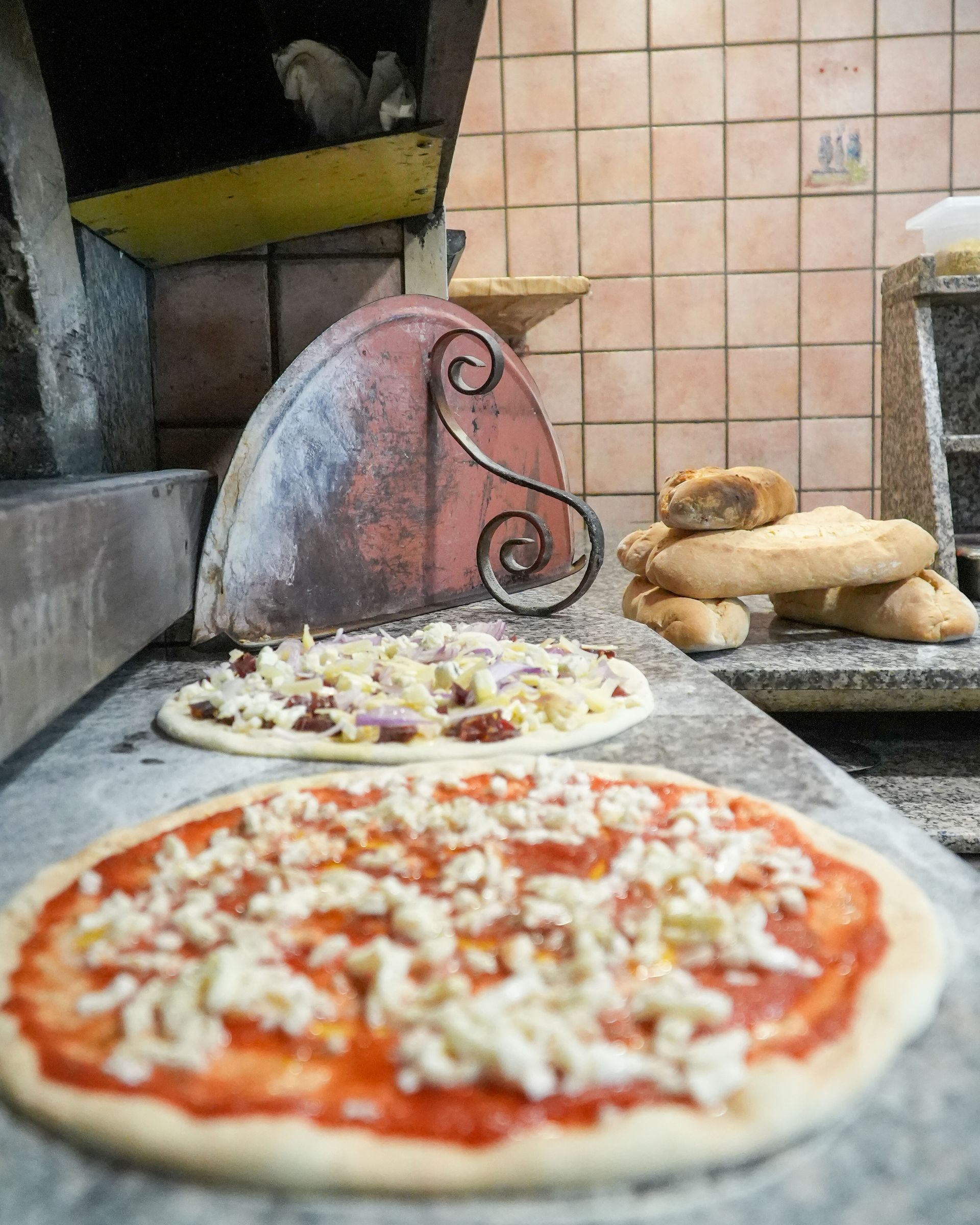 Le pizze vengono preparate in una cucina con forno a legna e rivestimento in piastrelle, con l'impasto e i condimenti disposti su un bancone di pietra.