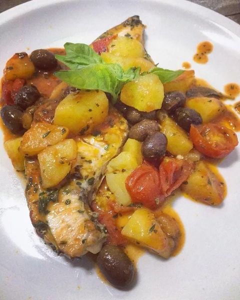 Pesce arrosto con patate, pomodorini e olive su un piatto bianco, guarnito con basilico.
