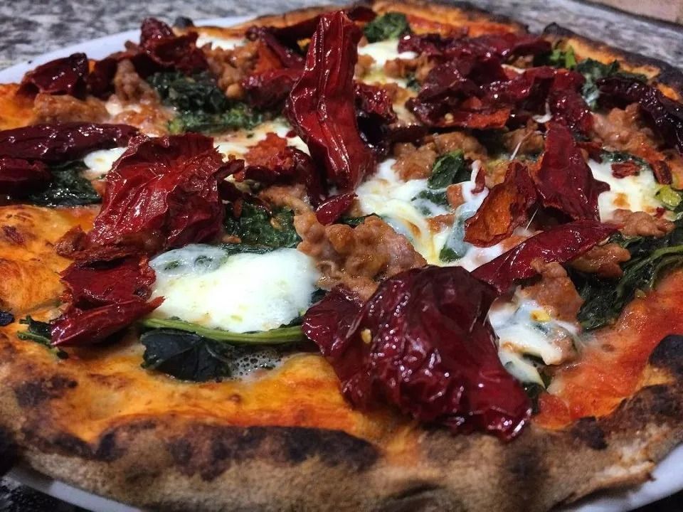Pizza condita con mozzarella, verdure appassite e pomodori secchi su una crosta bruciacchiata.