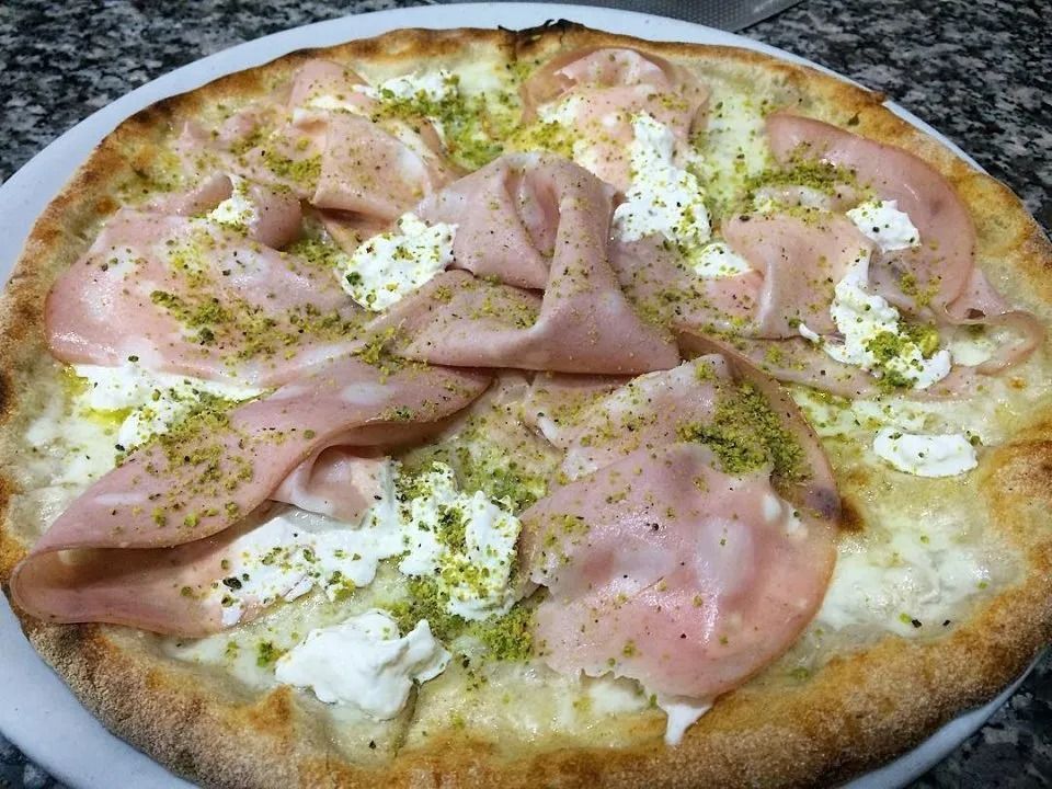 Pizza condita con prosciutto, formaggio cremoso ed erbe aromatiche su un piatto bianco.
