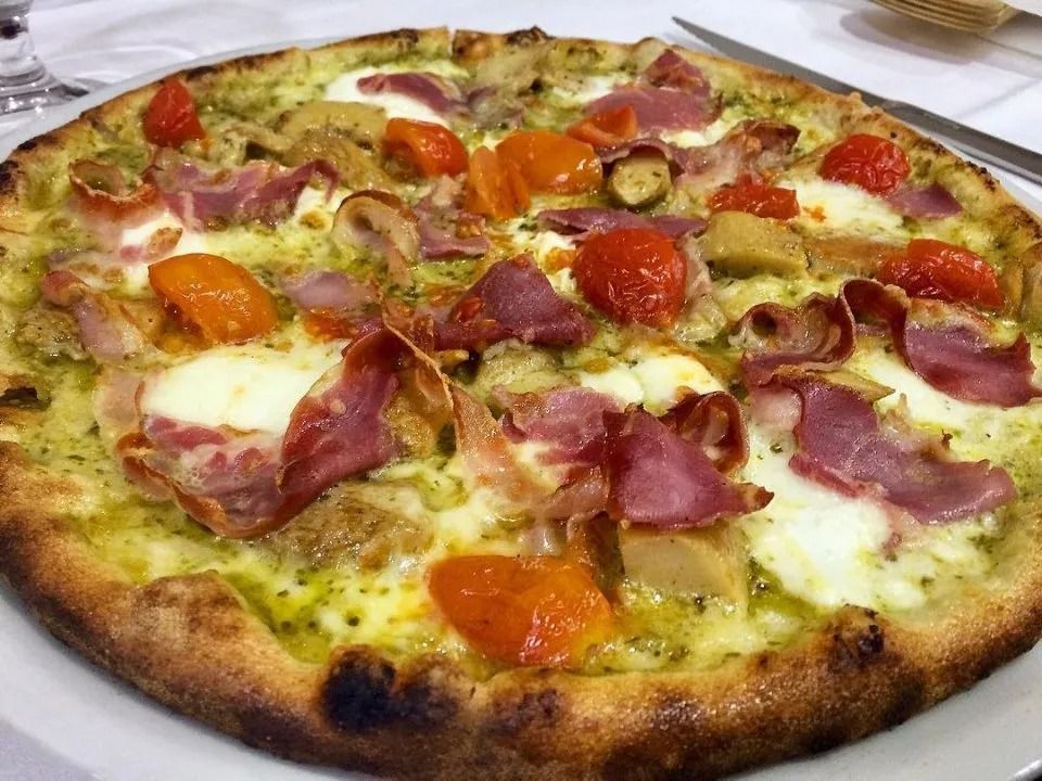 Primo piano di una pizza cotta al forno con formaggio fuso, fette di pomodoro, salumi e pesto verde su un piatto bianco.