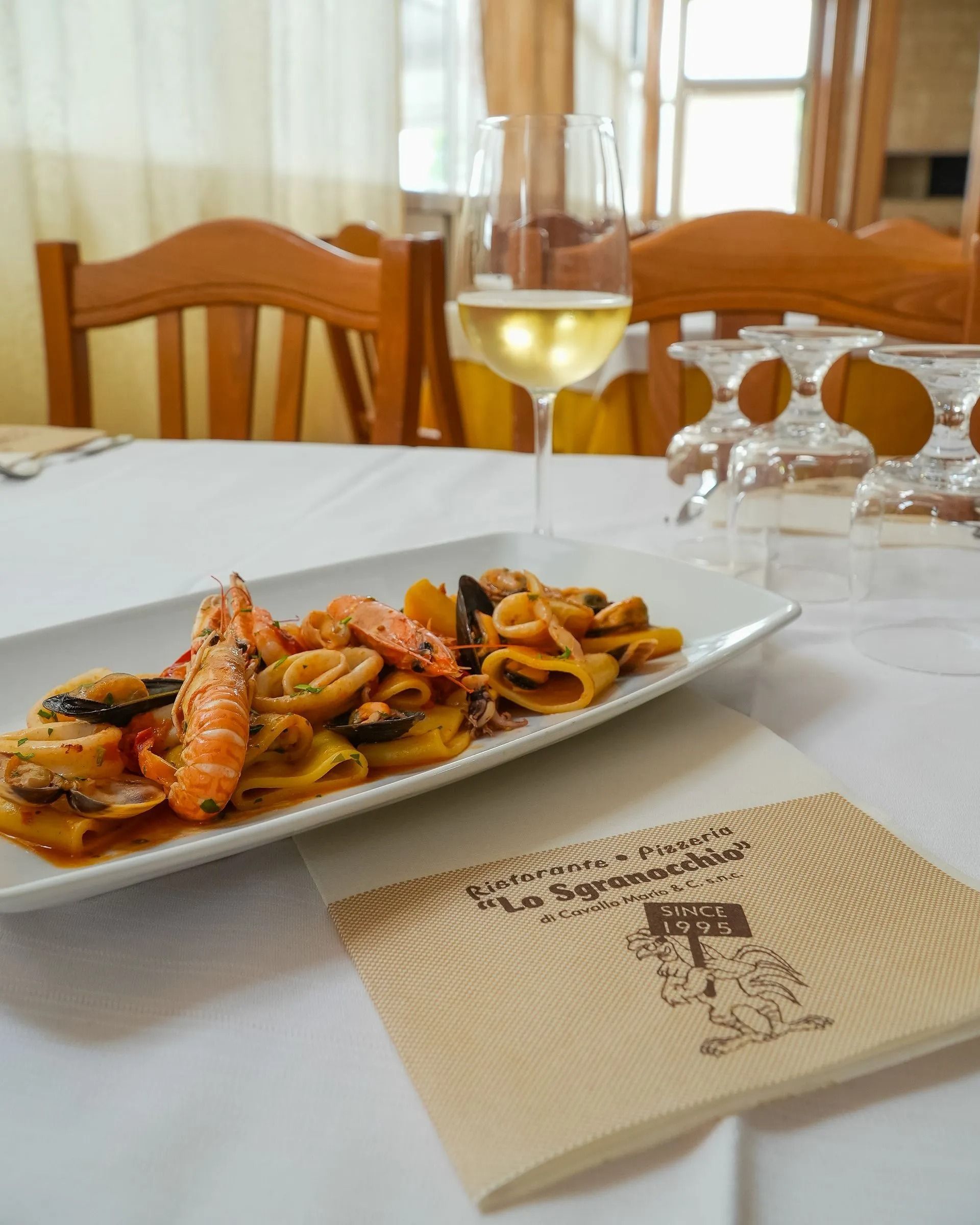 Plate of pasta and seafood on a white table beside a glass of white wine in a restaurant