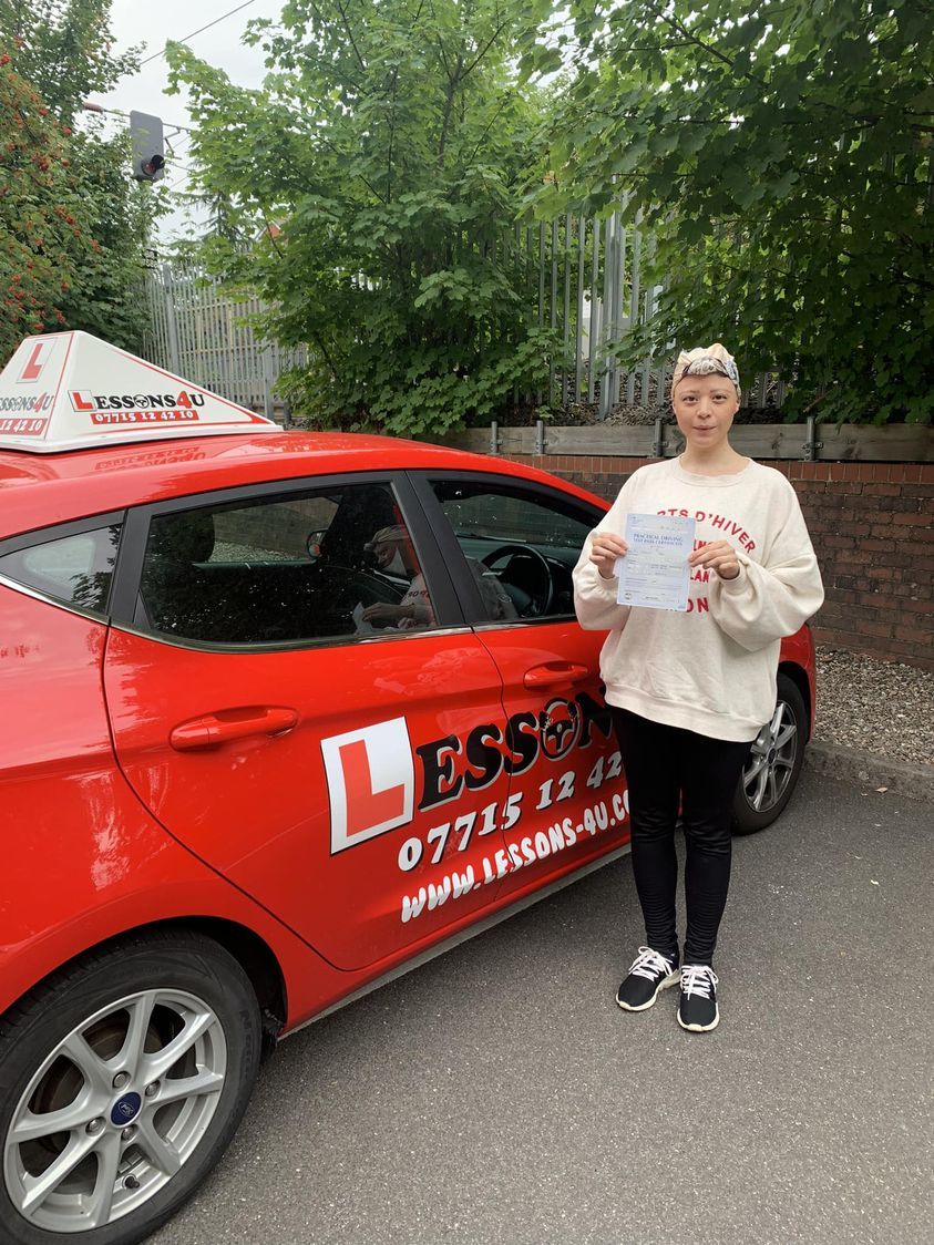 A person stands next to a red driving school car, holding a certificate to celebrate passing their driving test.