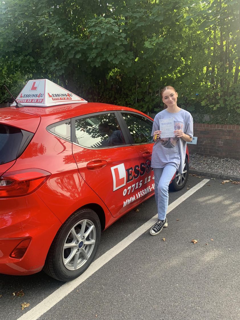 A person stands smiling beside a red driving school car with an L-sign on the roof, holding a driving test pass certificate.