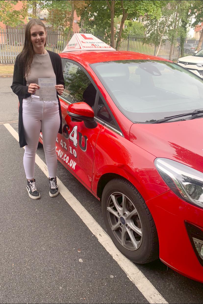 A person stands next to a red driving school car with an L-plate, smiling while holding a document.