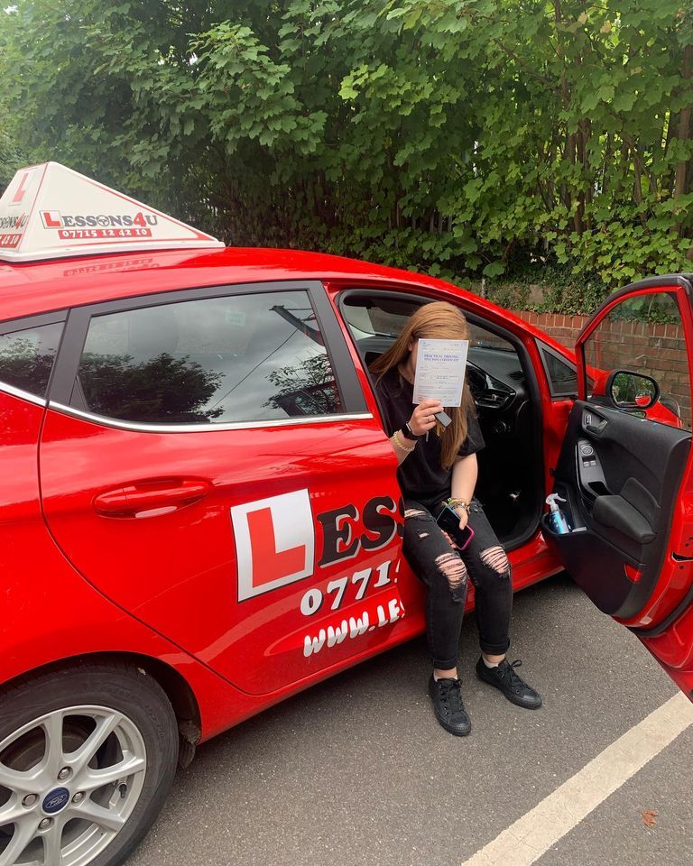 A person sitting in a bright red driving school car with an L-plate, holding a paper over their face.