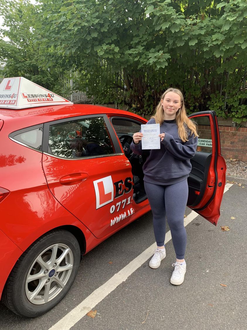 A smiling person stands next to a red driving school car holding a passed driving test certificate.