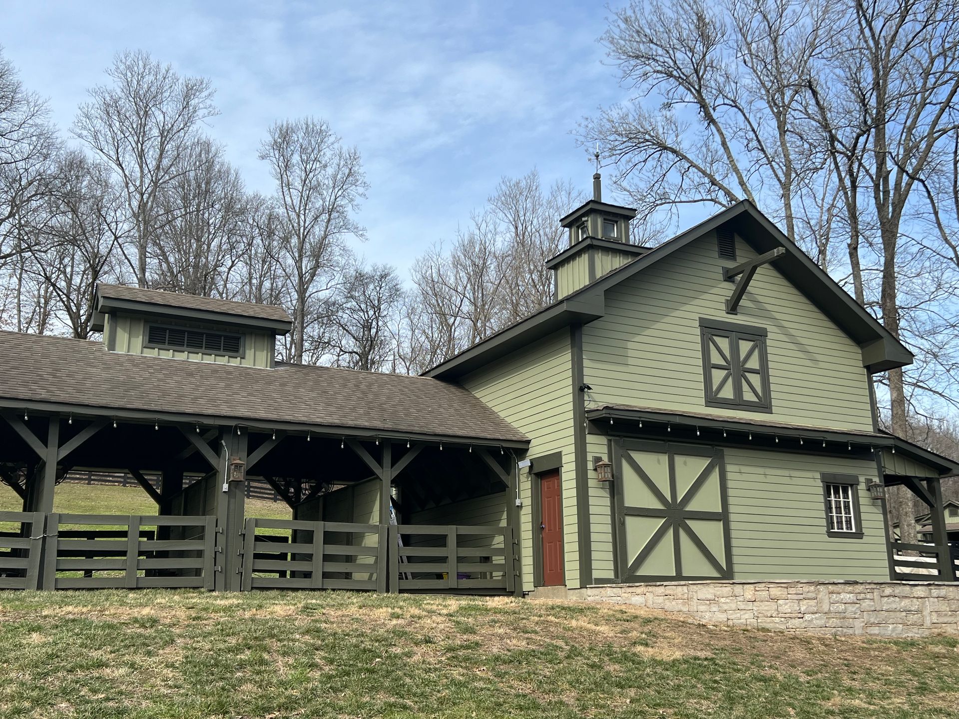 A green barn with a brown roof is sitting in the middle of a grassy field.