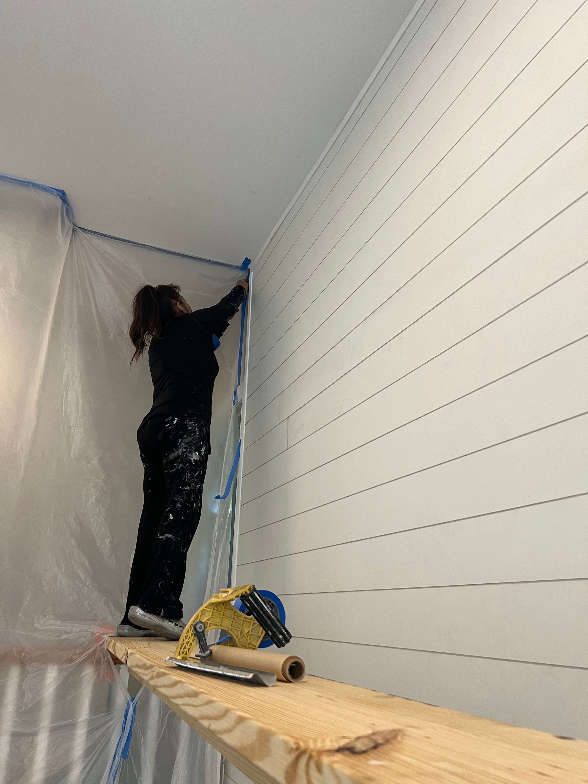 A woman is standing on a wooden shelf painting a wall.