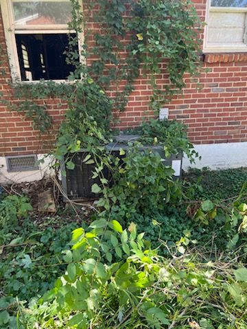 A brick house with a broken window and a air conditioner surrounded by plants.