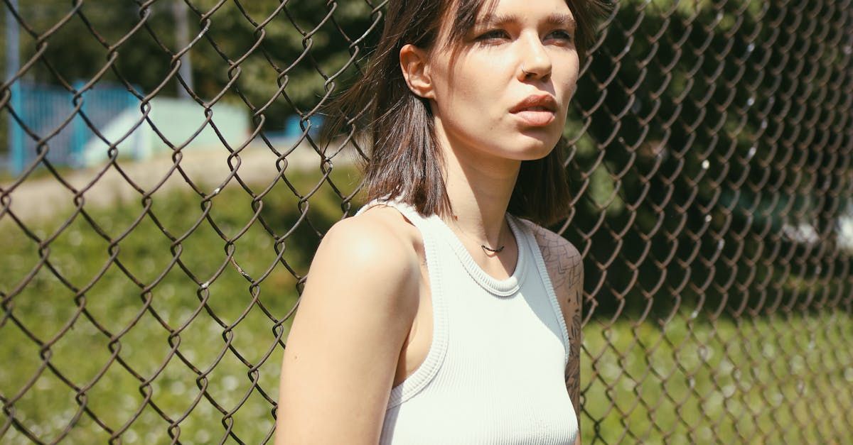 A woman in a white tank top is standing next to a chain link fence.