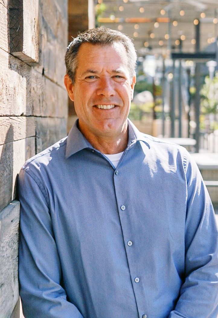 Man in a blue button-down shirt smiling beside a stone wall outdoors