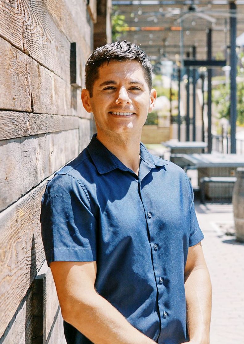 Man in a blue shirt leaning against a stone wall on a sunny sidewalk
