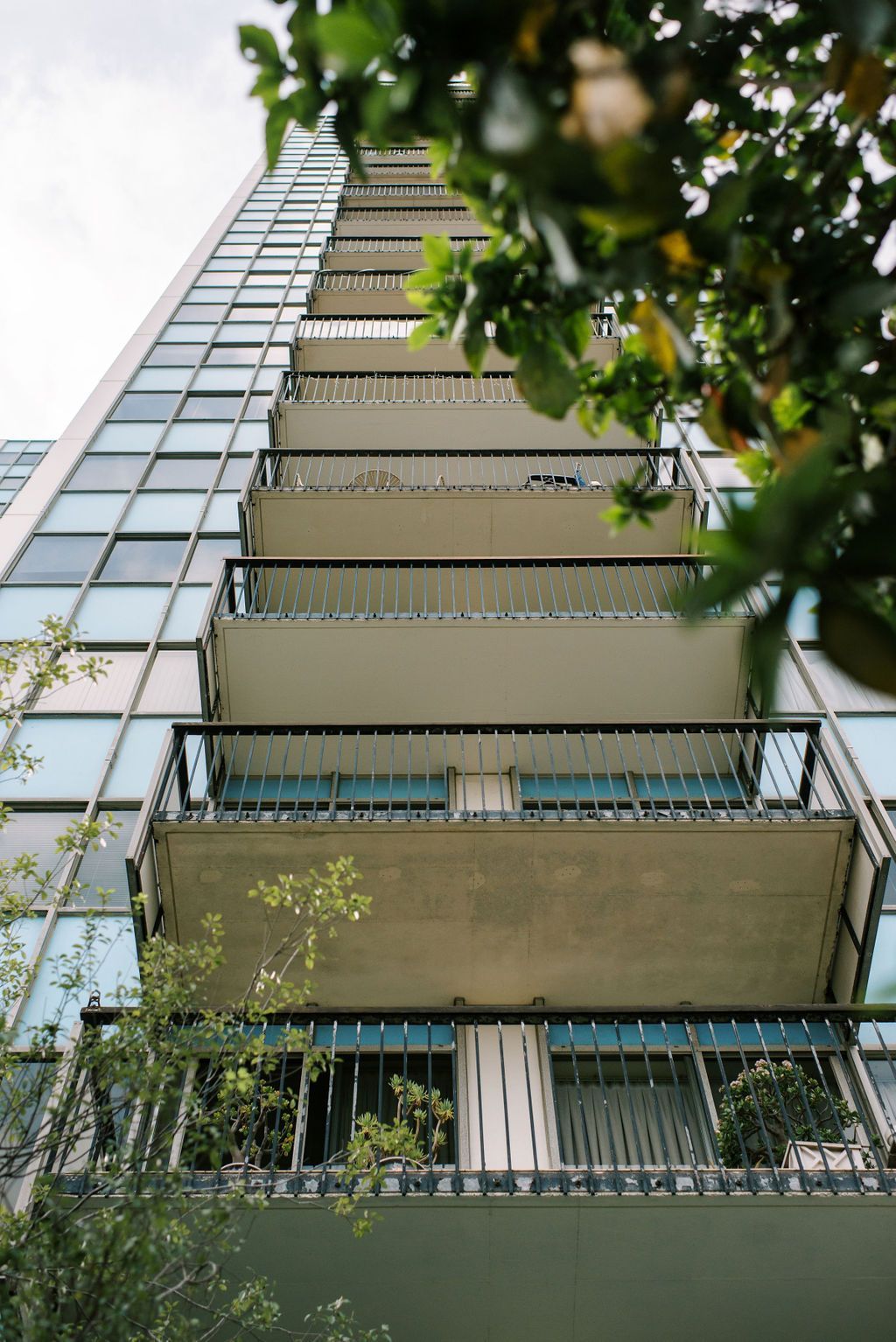 Looking up at a tall building with balconies and trees in the foreground.