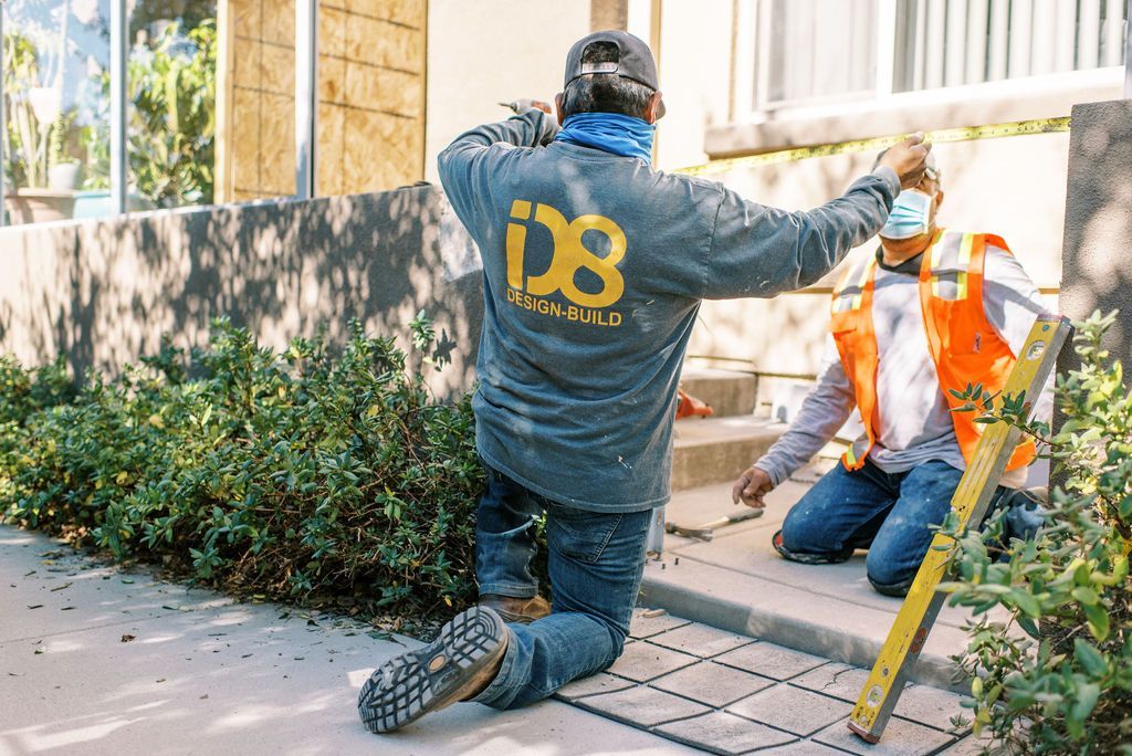 A man is kneeling down on the ground while another man paints a wall.