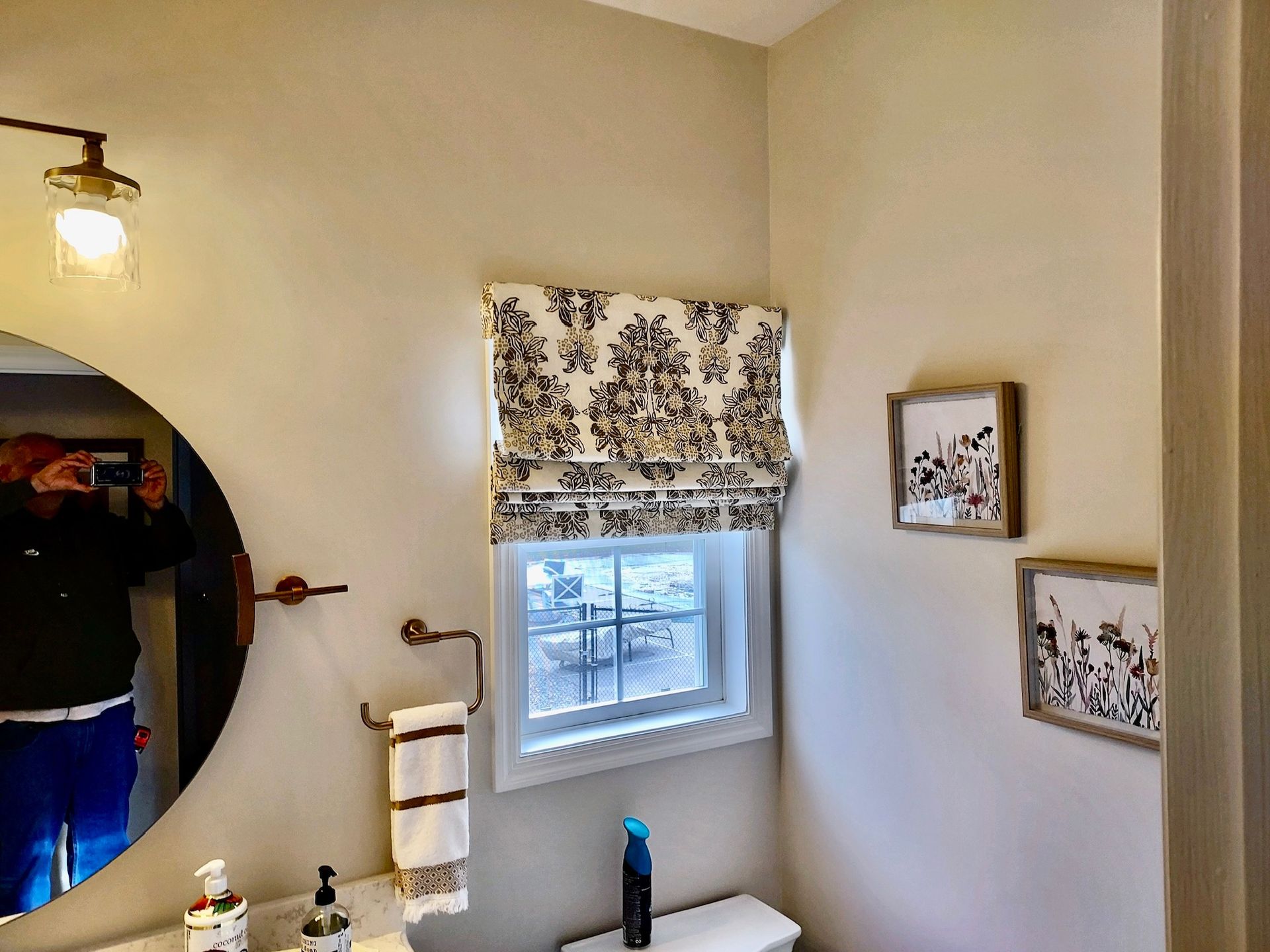 Bathroom with a round mirror, window with patterned shade, and framed art.