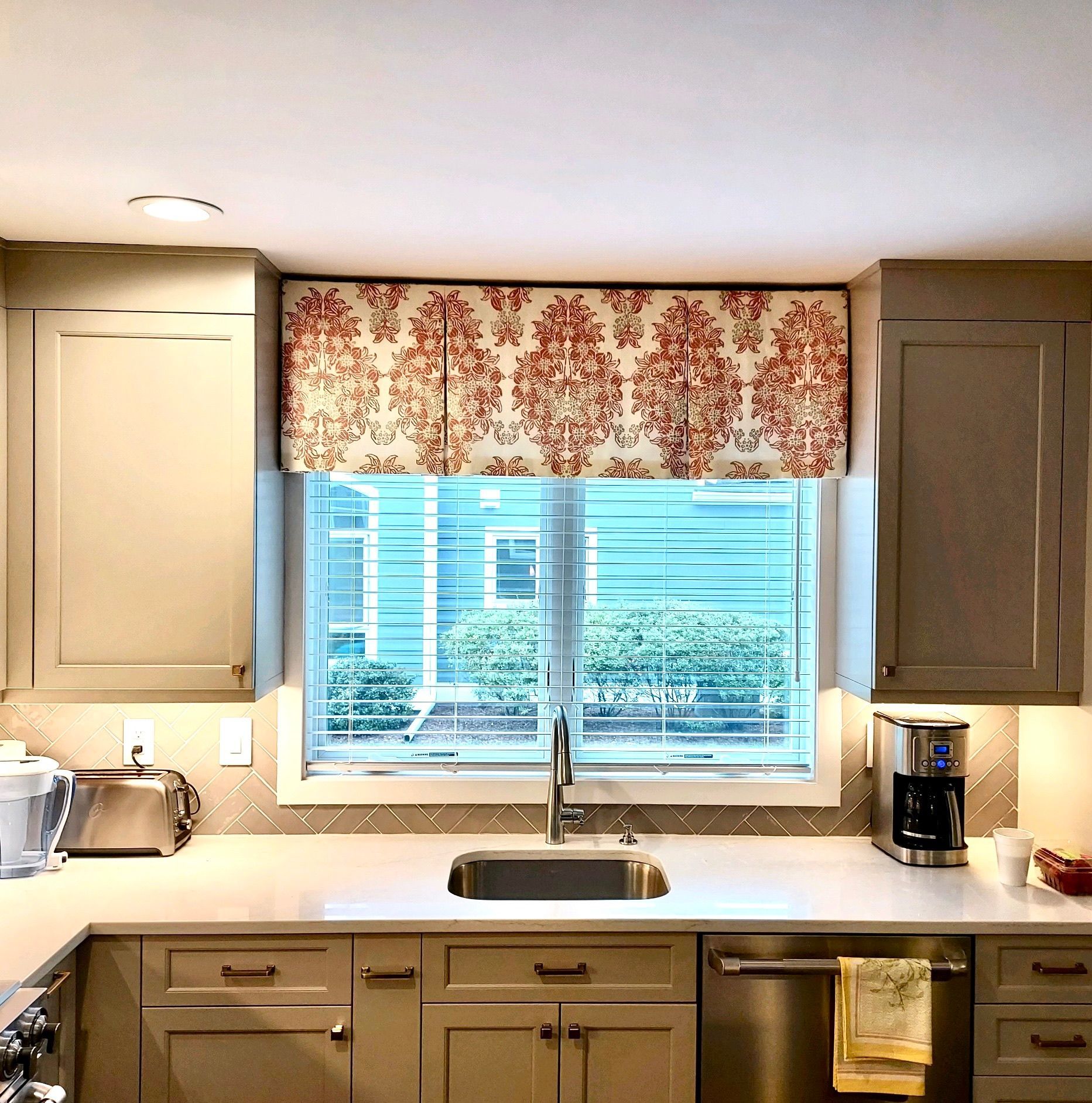 Kitchen with floral valance, window, sink, cabinets, appliances; neutral tones, natural light.