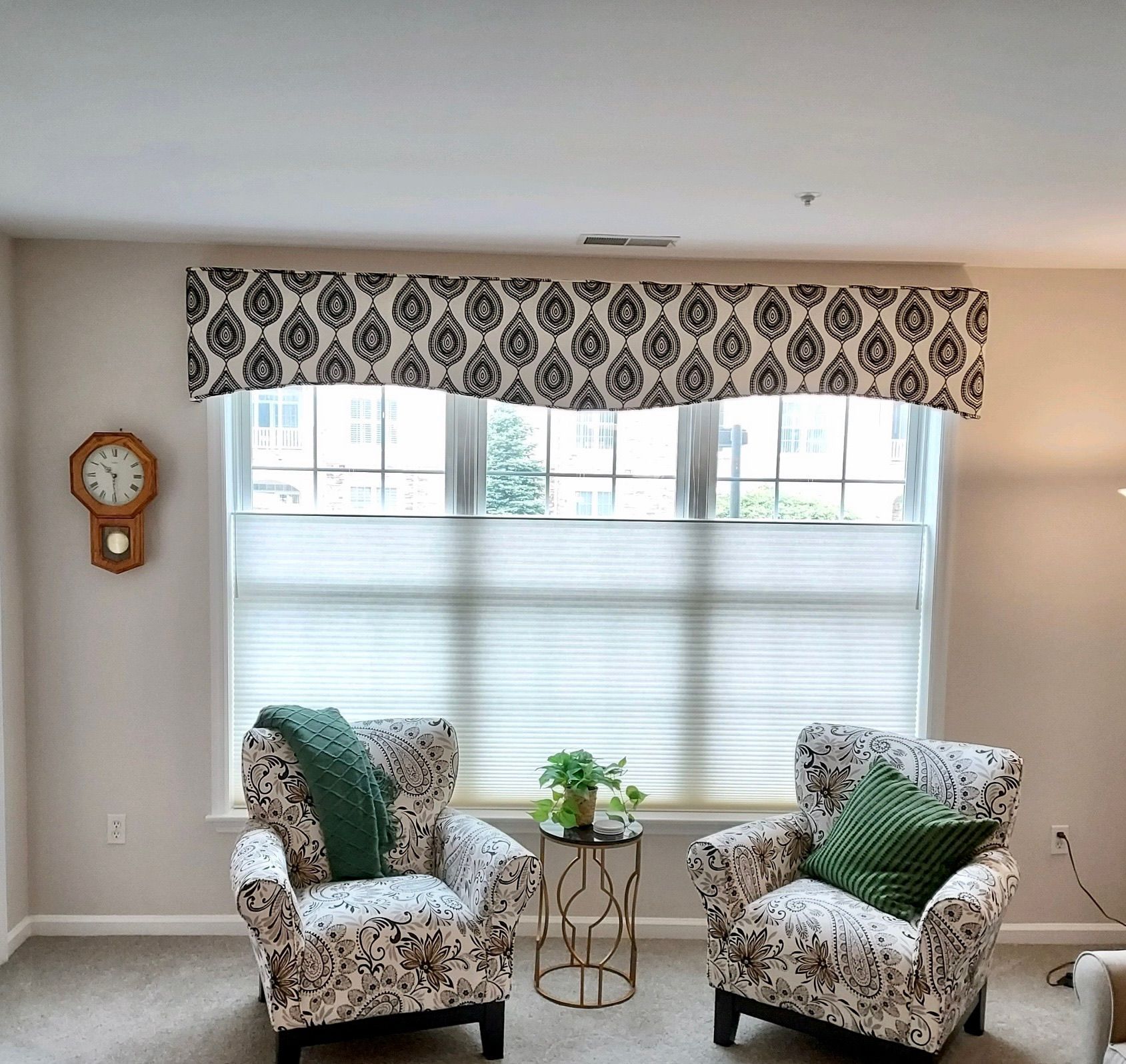 Two patterned armchairs face a window with a patterned valance and pleated shades. A small table and clock are nearby.