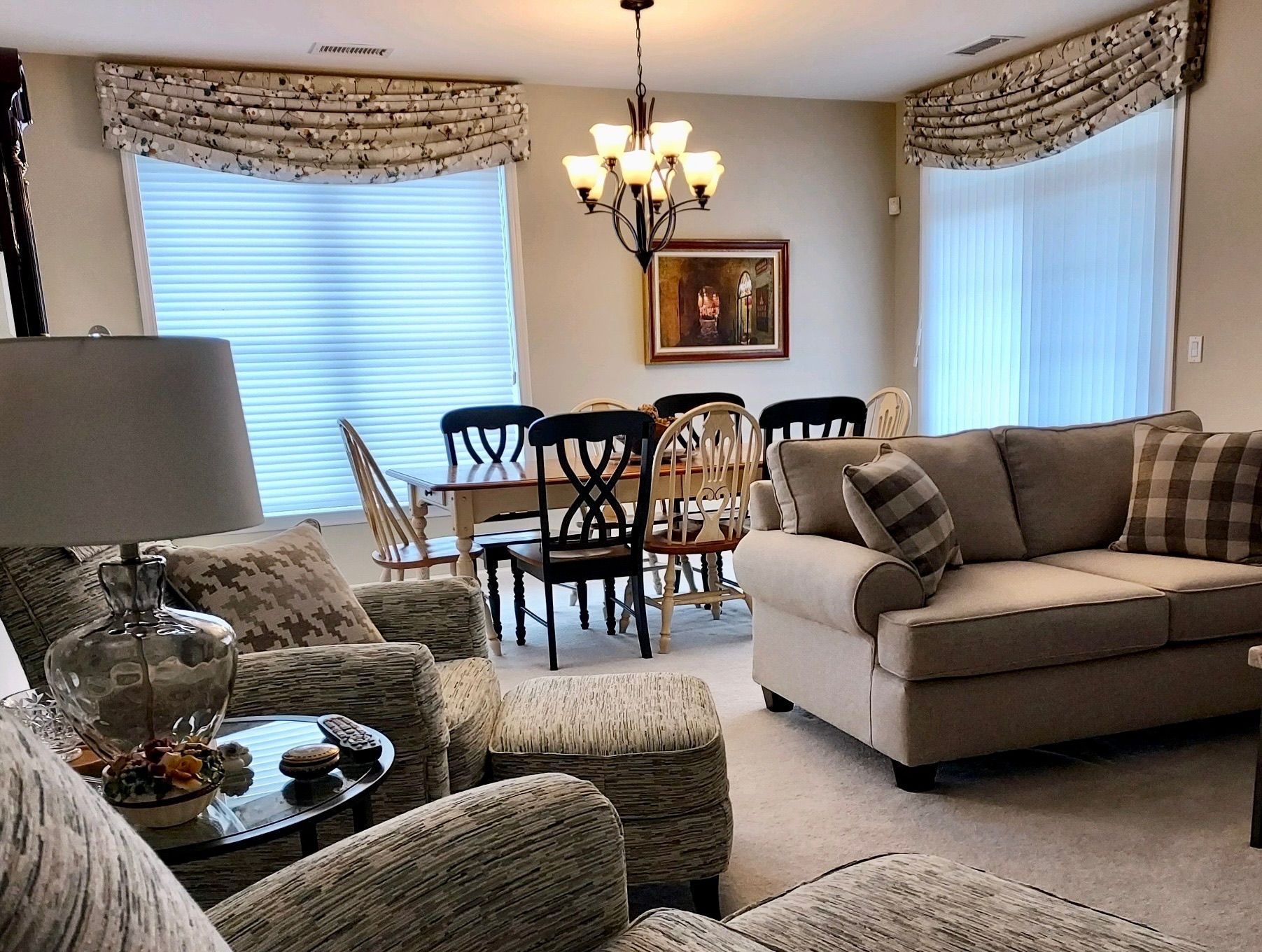 Living room with neutral tones: sofa, armchair, dining table, chandelier, window treatments.