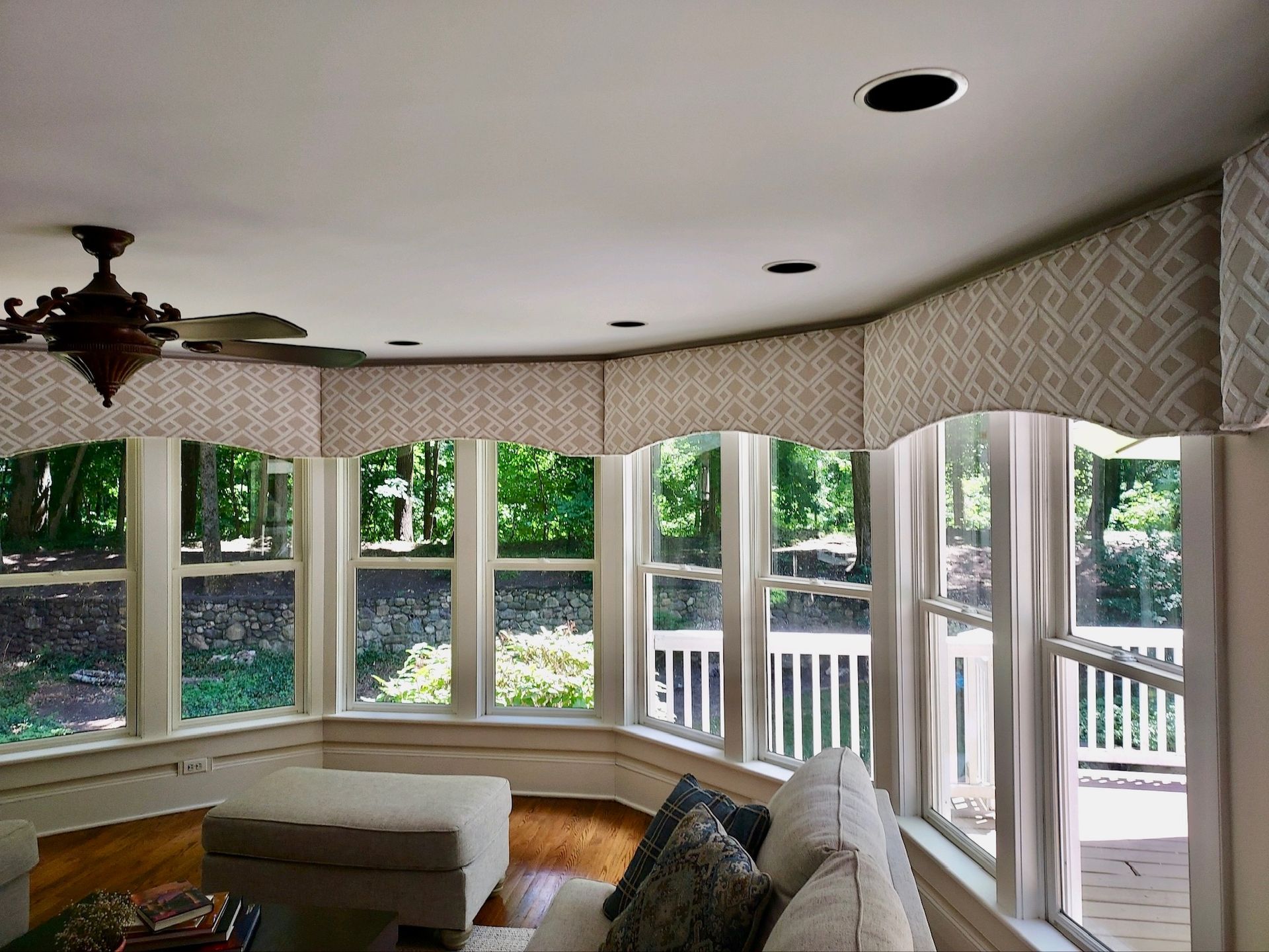 A sunroom with large windows, white trim, and beige geometric valances. A white sofa and ottoman sit inside.