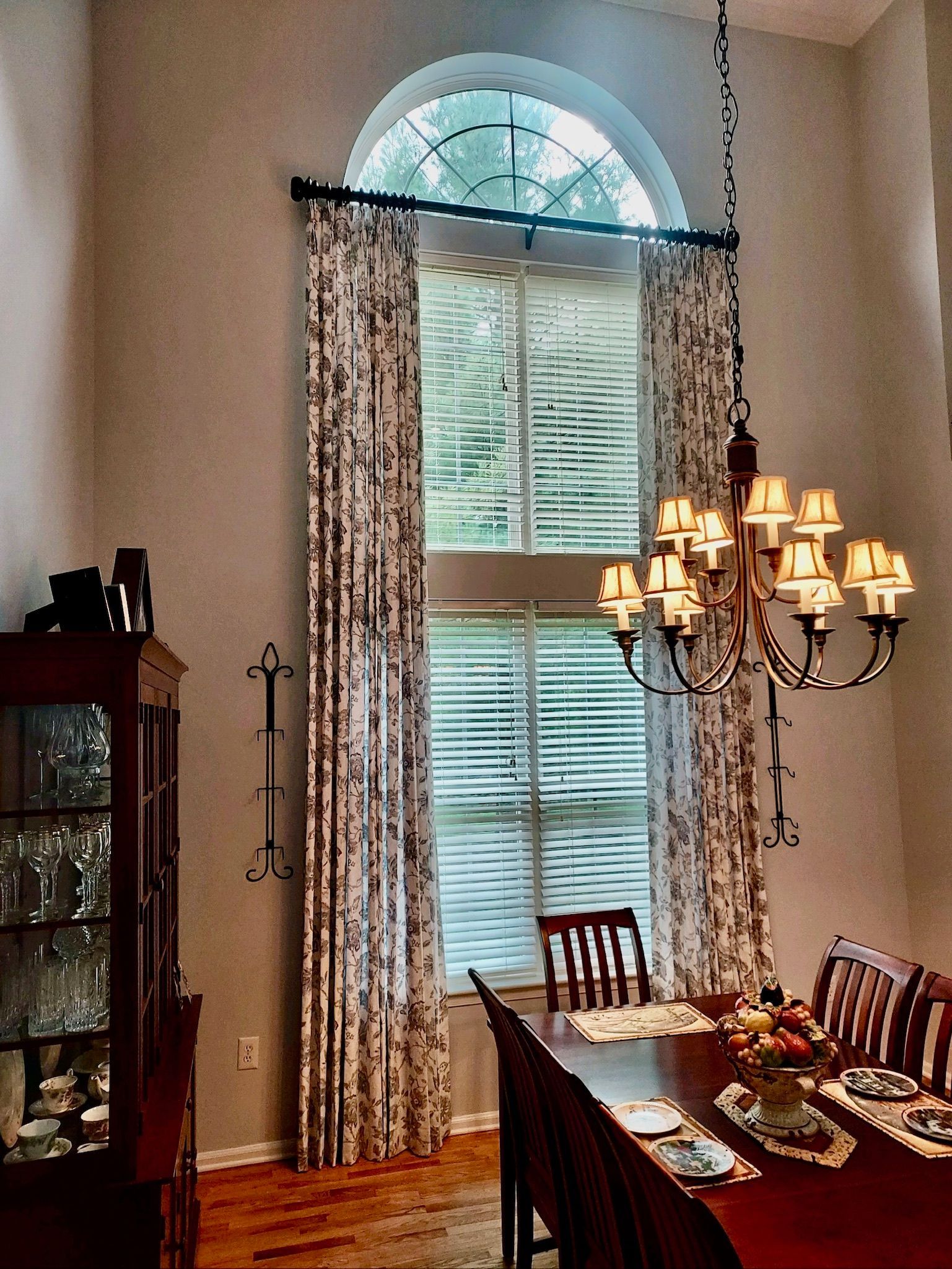 Dining room with large arched window, patterned curtains, dining table, and chandelier.