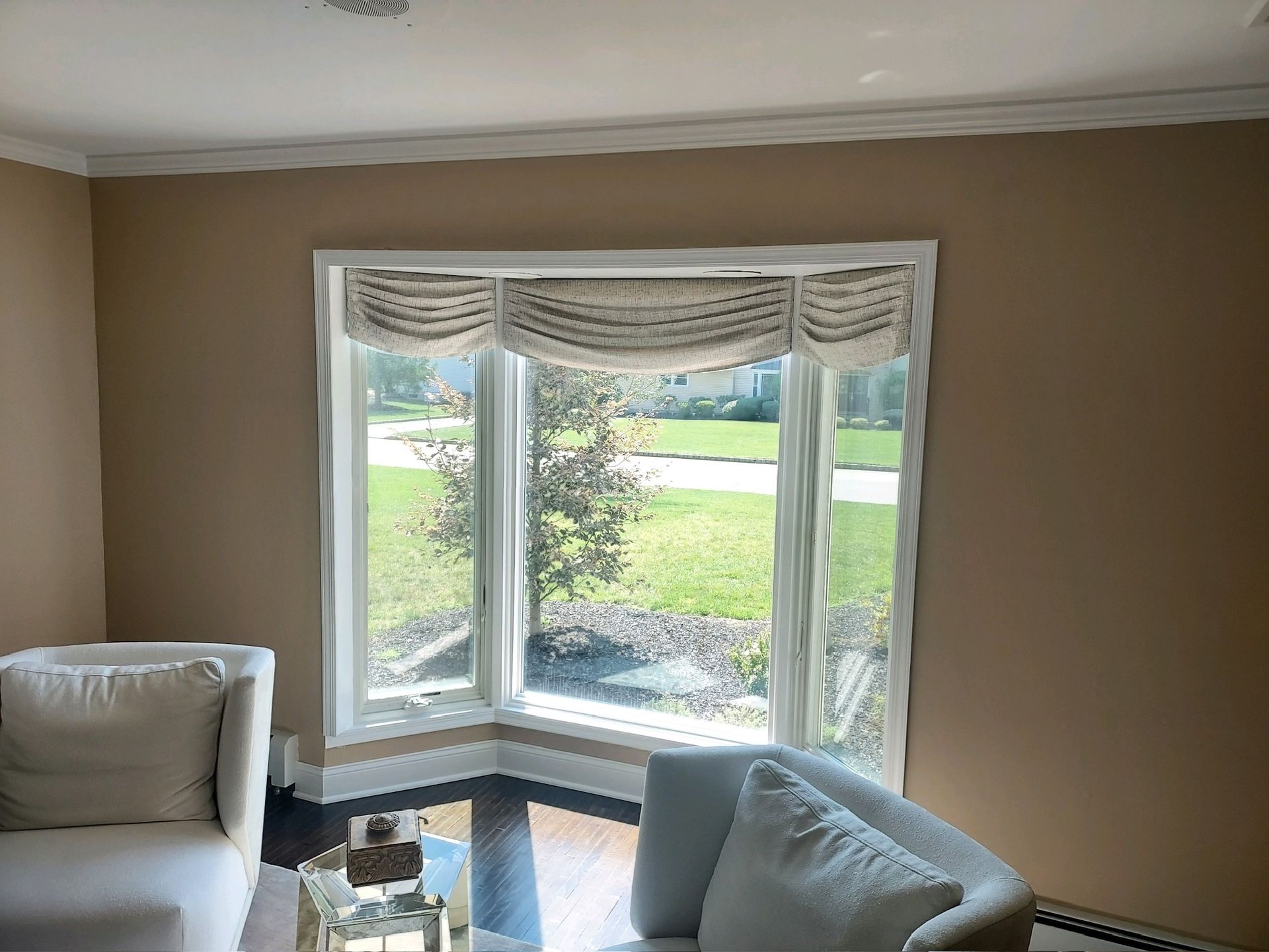 A bay window with a patterned shade, facing a yard, between beige walls. Two white armchairs are in the corner.