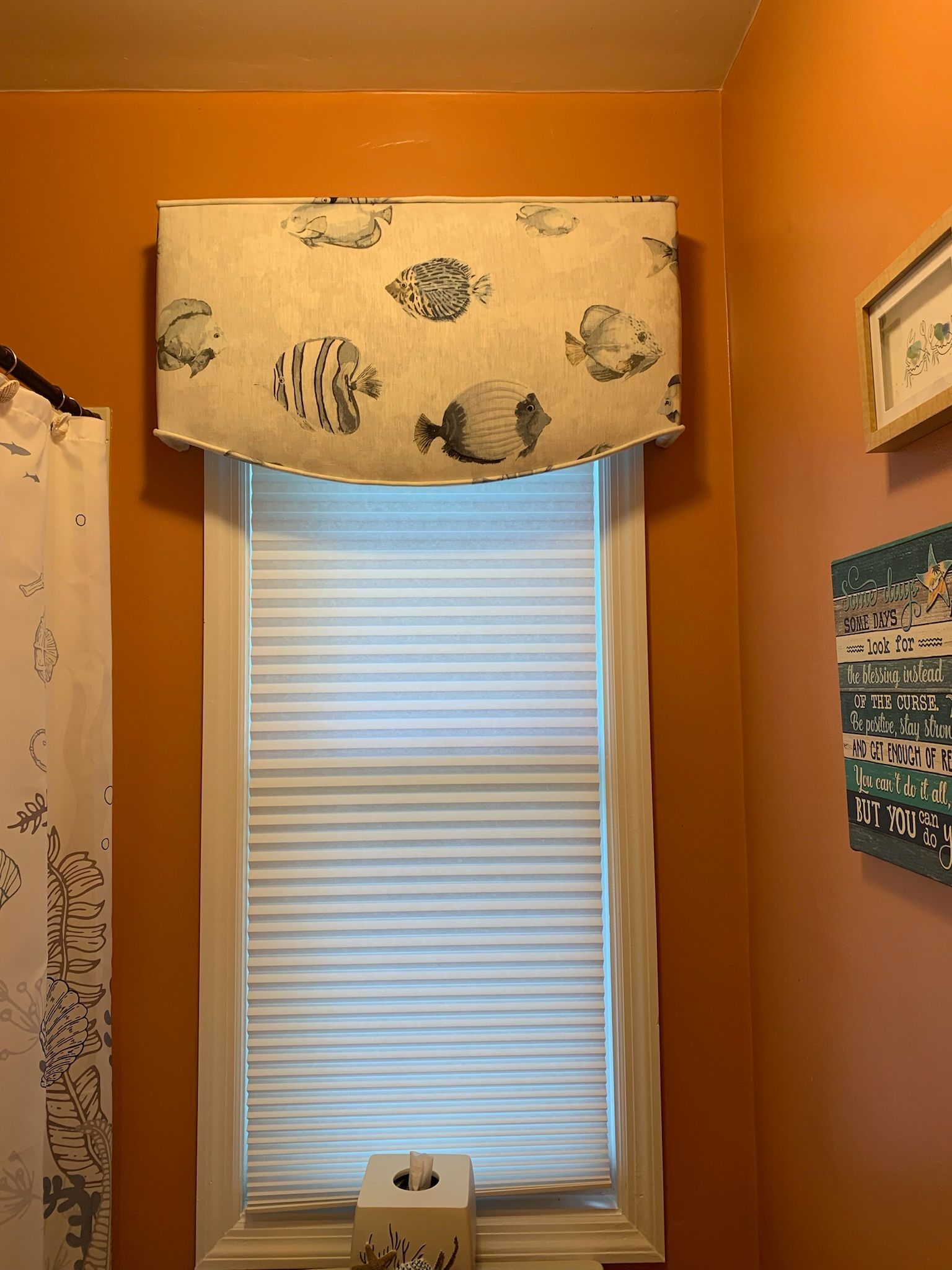 Window with a fish-patterned valance and white blinds in a bathroom with orange walls.