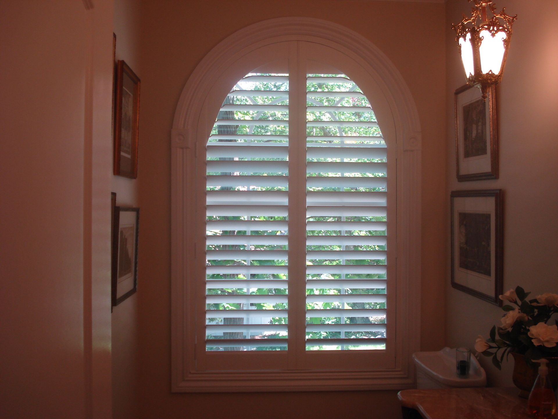 Arched window with white shutters, framed with ornate molding. Interior room with artwork and lamp.