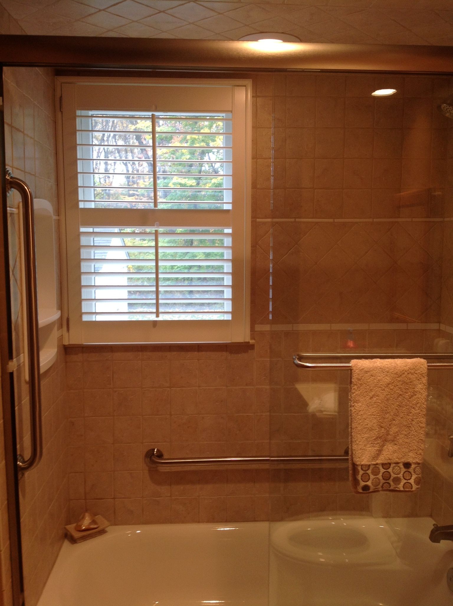 Bathroom with shower, tub, window with shutters, and grab bars. Neutral tiles and beige towel.