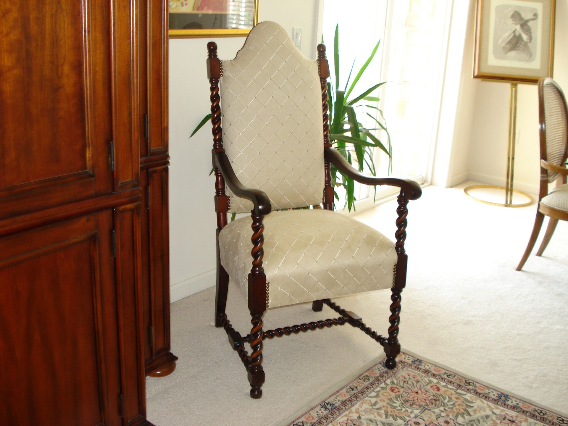Wooden armchair with patterned beige upholstery, in a room with a tall plant and framed artwork.